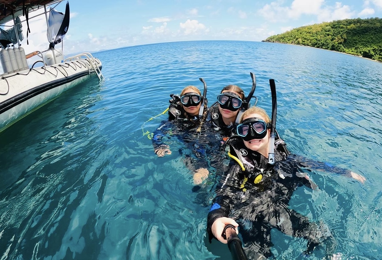 Three students in scuba gear using a selfie stick to take a photo while in the water. Behind them is a boat, a large body of blue water that they are resting in, and a dense green coastline.