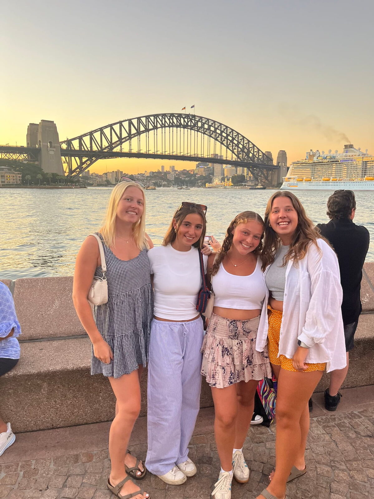 Four students posing together along the water at sunset. A large metal bridge can be seen behind them.