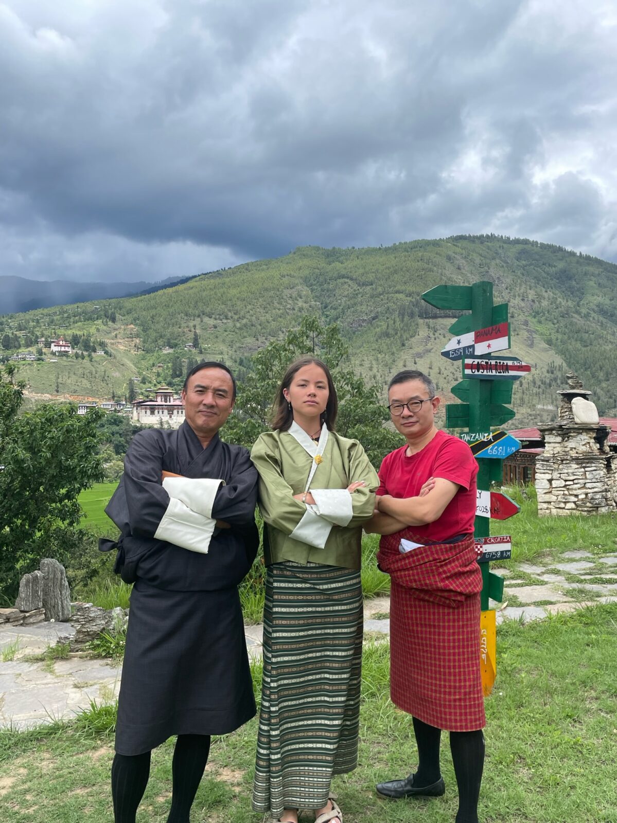 Three people in traditional clothing, posing with their arms crossed. Behind them is a painted wooden tower with many arrows pointed in different directions, a green mountain with a couple houses nestled among the paths, and gray cloud cover.