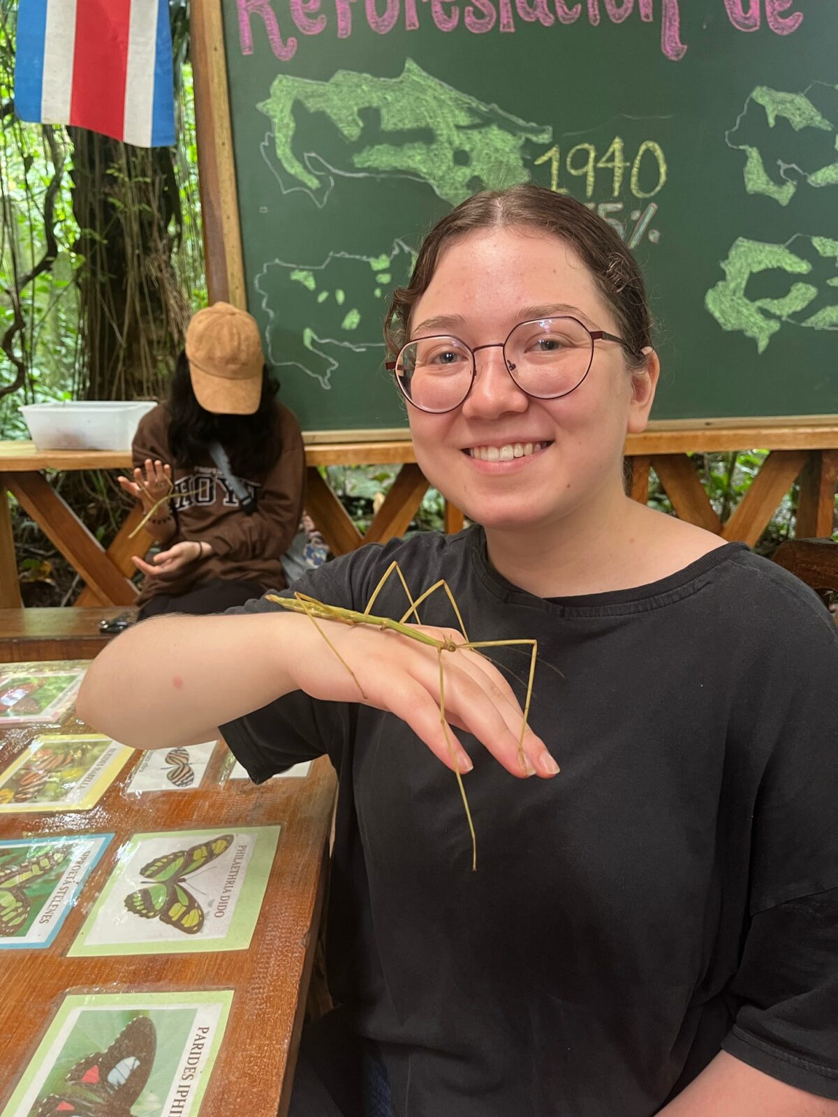 Adria sits at a table in an outdoor classroom while a Walking Stick perches on the back of her hand.