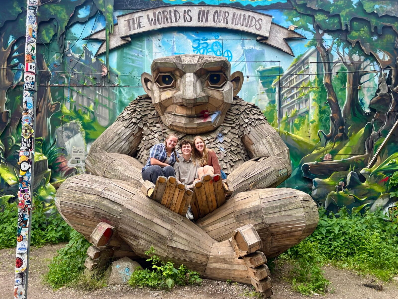 Three people smile and pose on a giant wooden statue of a troll sitting cross-legged in bushes.