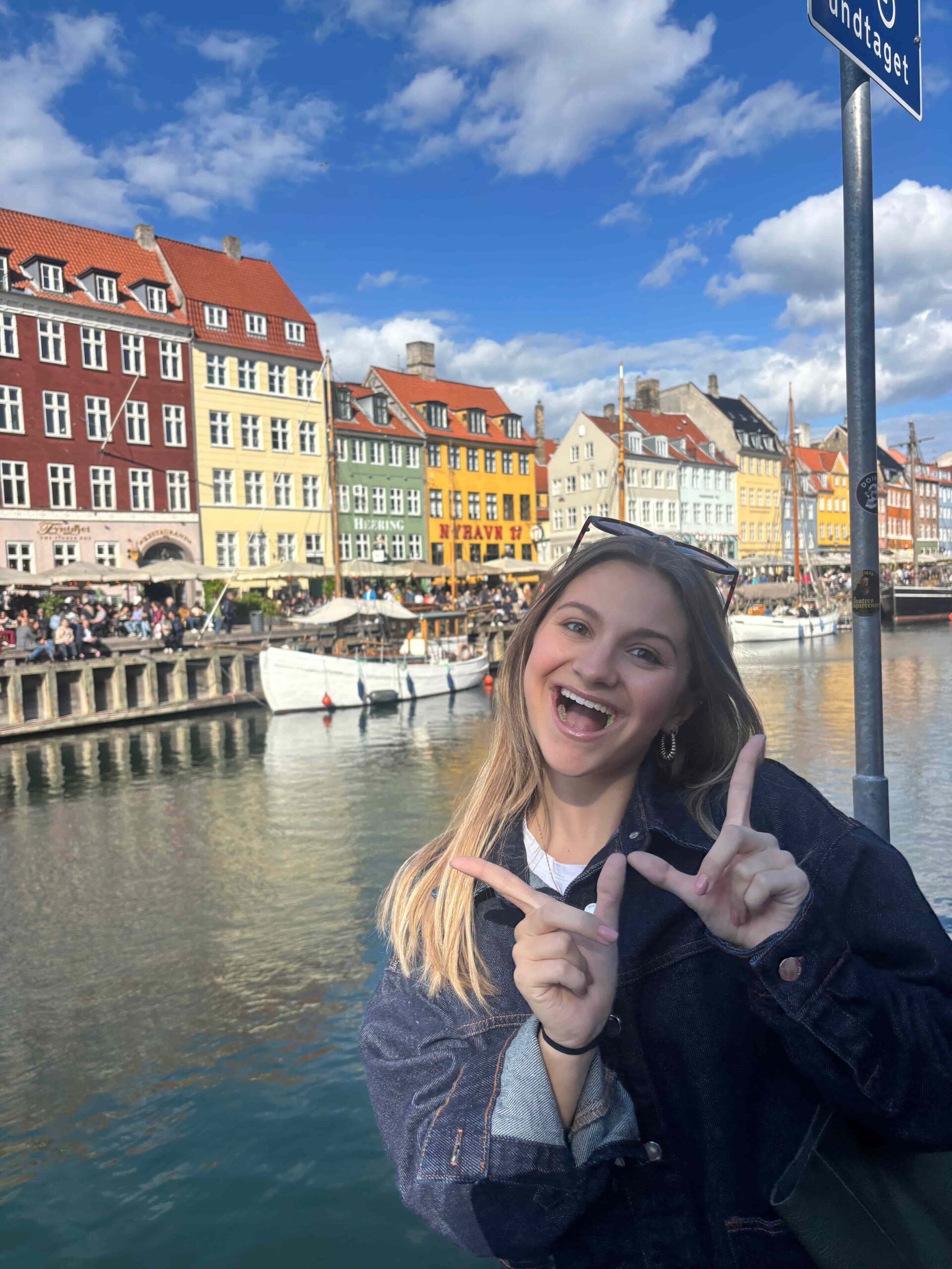 Student with long blonde hair makes a "W" with her fingers while standing in front of a canal. In the background are colorful buildings and a blue sky.