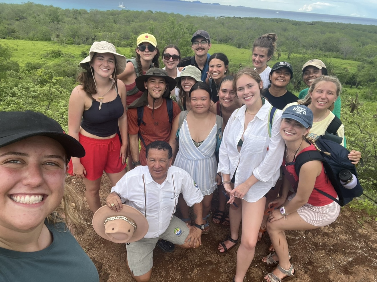 A group of students posed for a selfie with their guide in a grassy area overlooking distant forests.