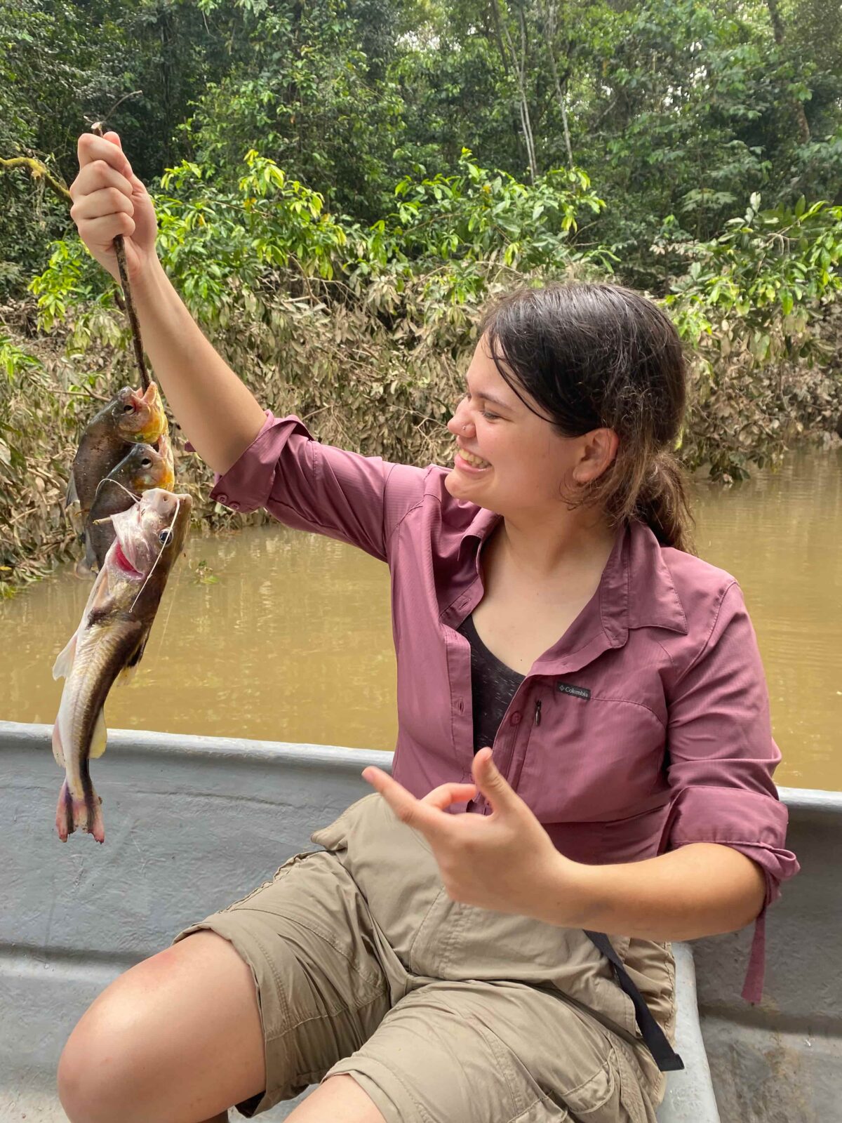 A student holds up a fish in her hand and smiles at the fish, pointing with her other hand. She is on a boat in a dirty river.