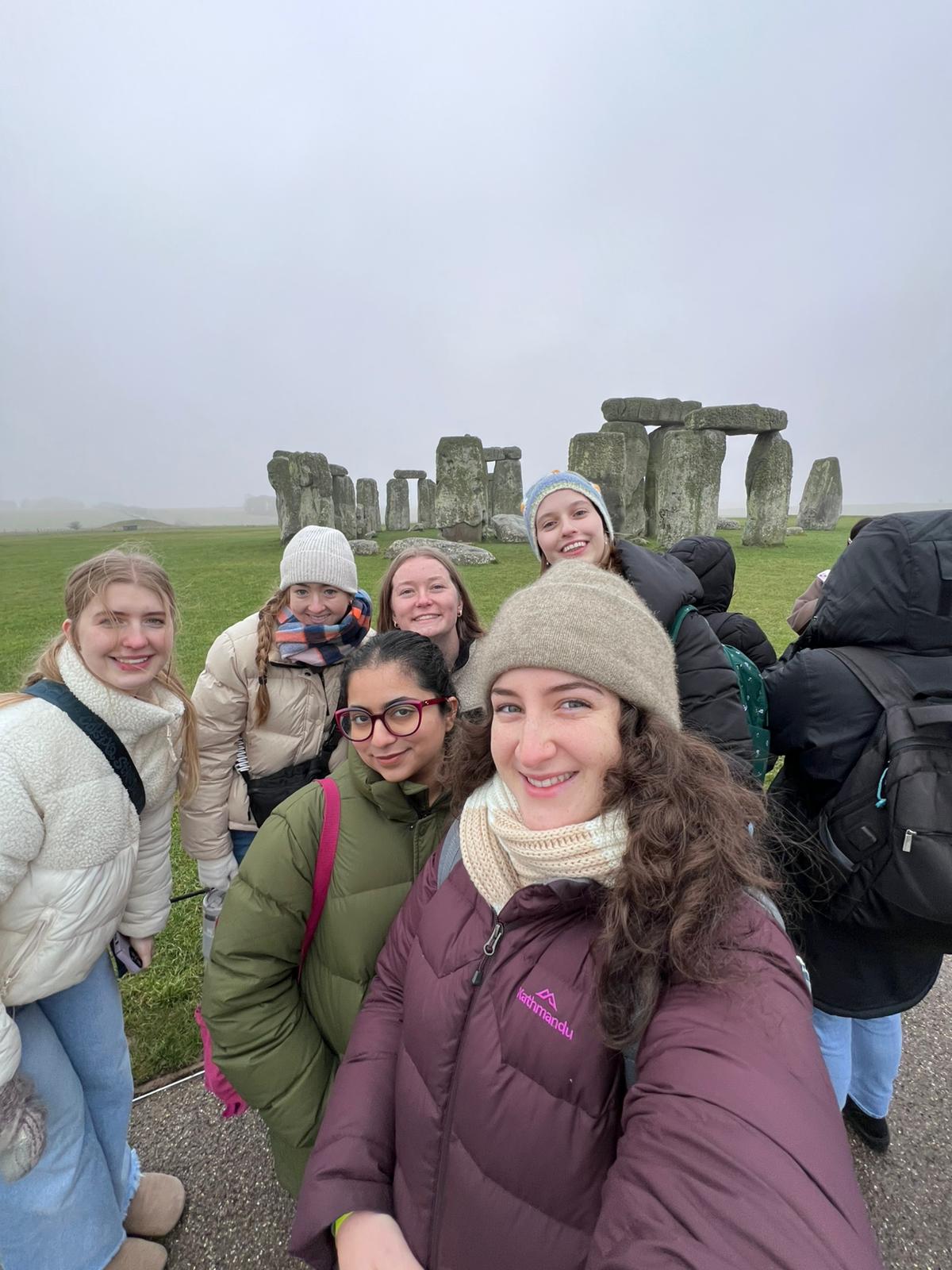 6 students are gathered for a selfie in front of the Stonehenge landmark on a foggy day. They are wearing winter jackets and hats.