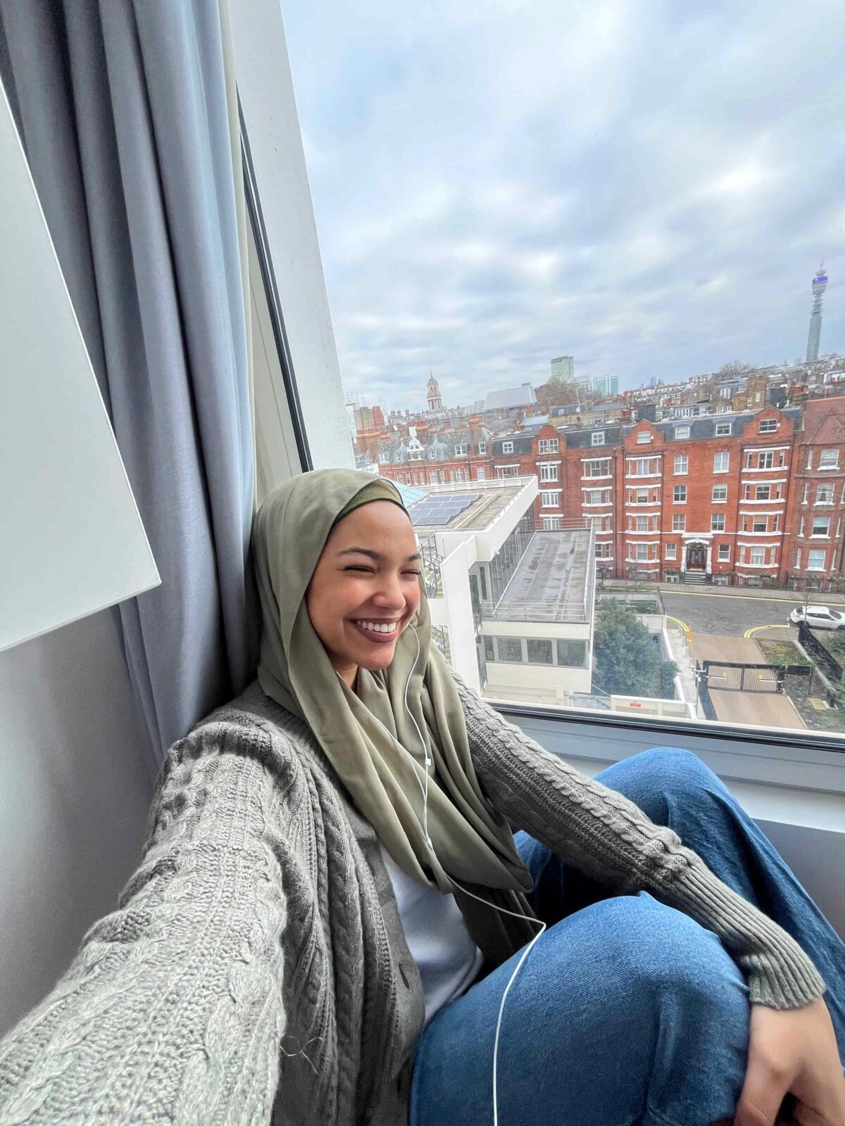 A selfie of a student wearing a headscarf. She is sitting next to her bedroom window and red brick buildings are visible outside.