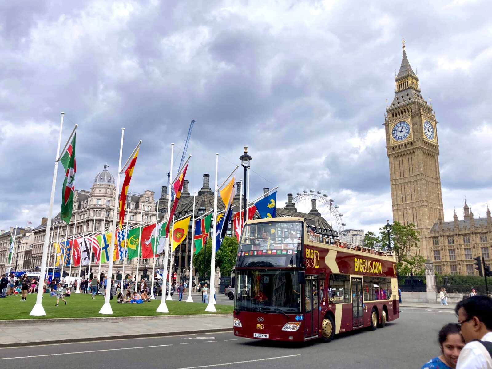 Flags of many different countries are mounted on poles on the edge of a grassy round-a-bout. A large, dark red double-decker bus drives by. The London Eye and Big Ben are seen in the back.