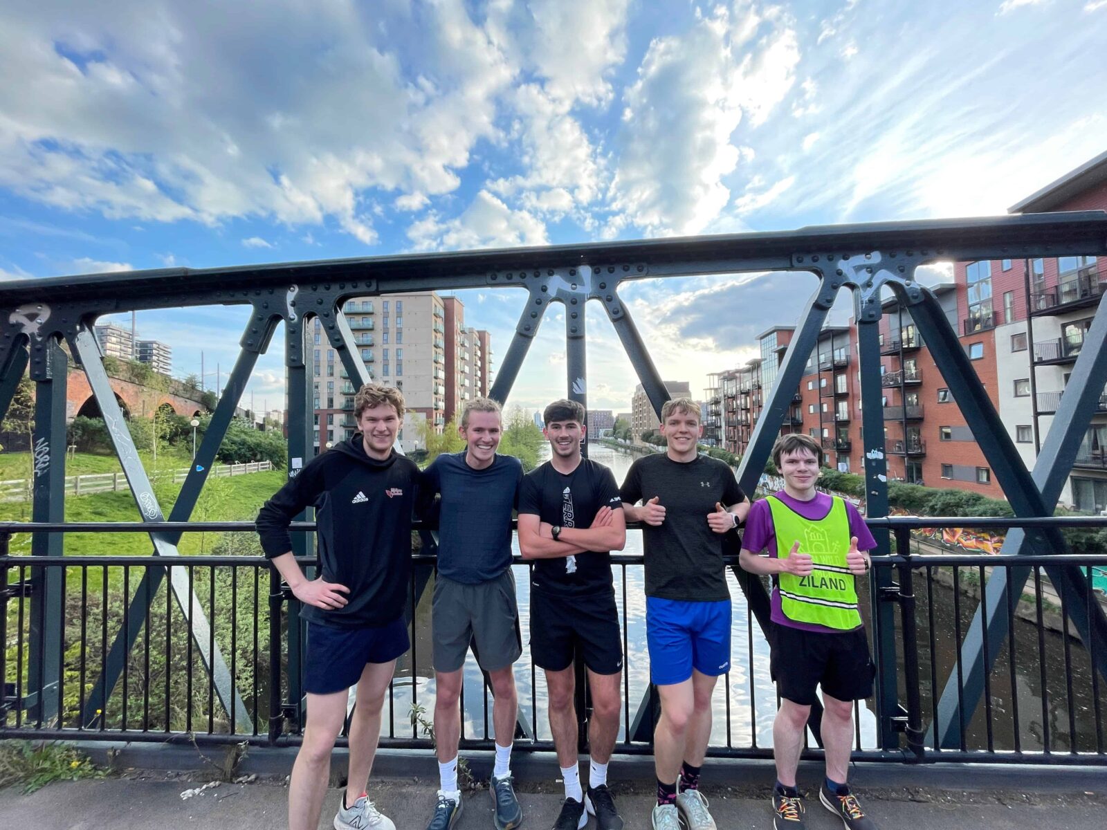 Five students are standing on a bridge over a waterway. They have just finished a run, and are in athletic clothes. The students are posed for the photo and smiling for the camera.