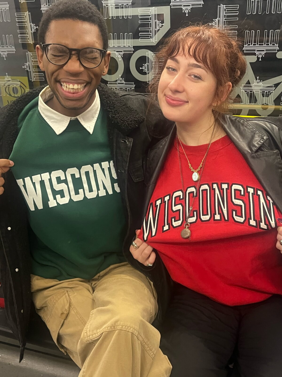 Two students holding open their jackets to show their green and red Wisconsin sweatshirts.