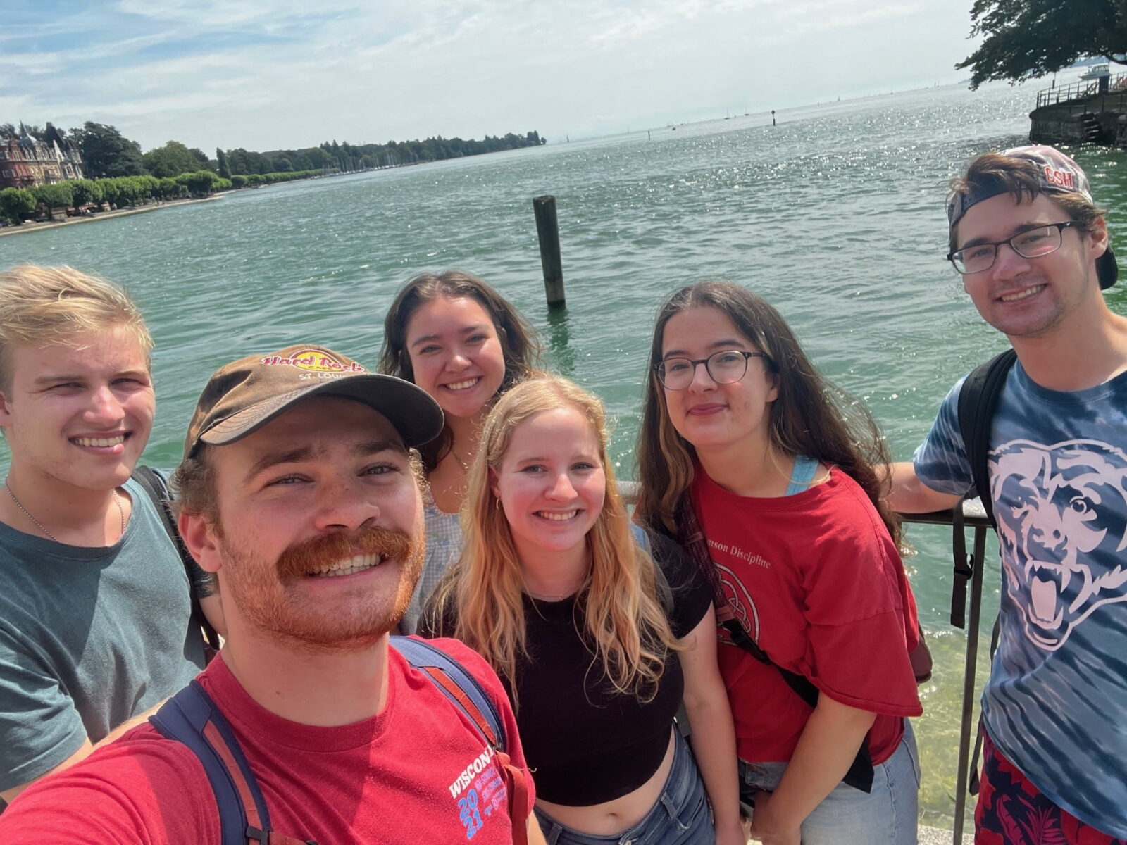 Six students posing for a selfie at the edge of a large lake.