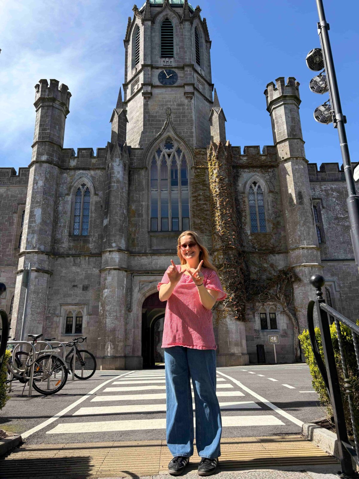 Kayla makes a "W" with her hands in front of a gothic stone building with arching stain glass windows and a large center turret.