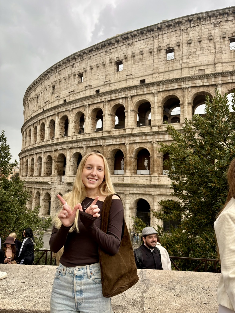 A student making a "W" with their fingers, while standing in the front of the Colosseum.
