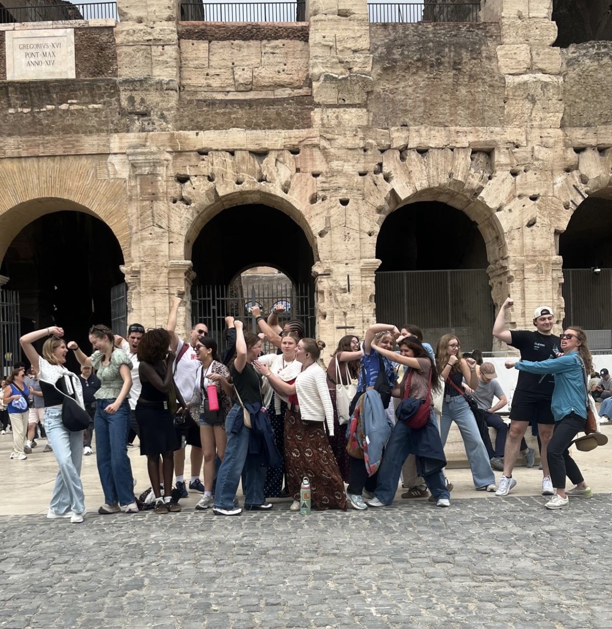 A large group of students clustered together pretend to be gladiators, paused in combat while holding imaginary swords. In the background are stone arches of a historical building.