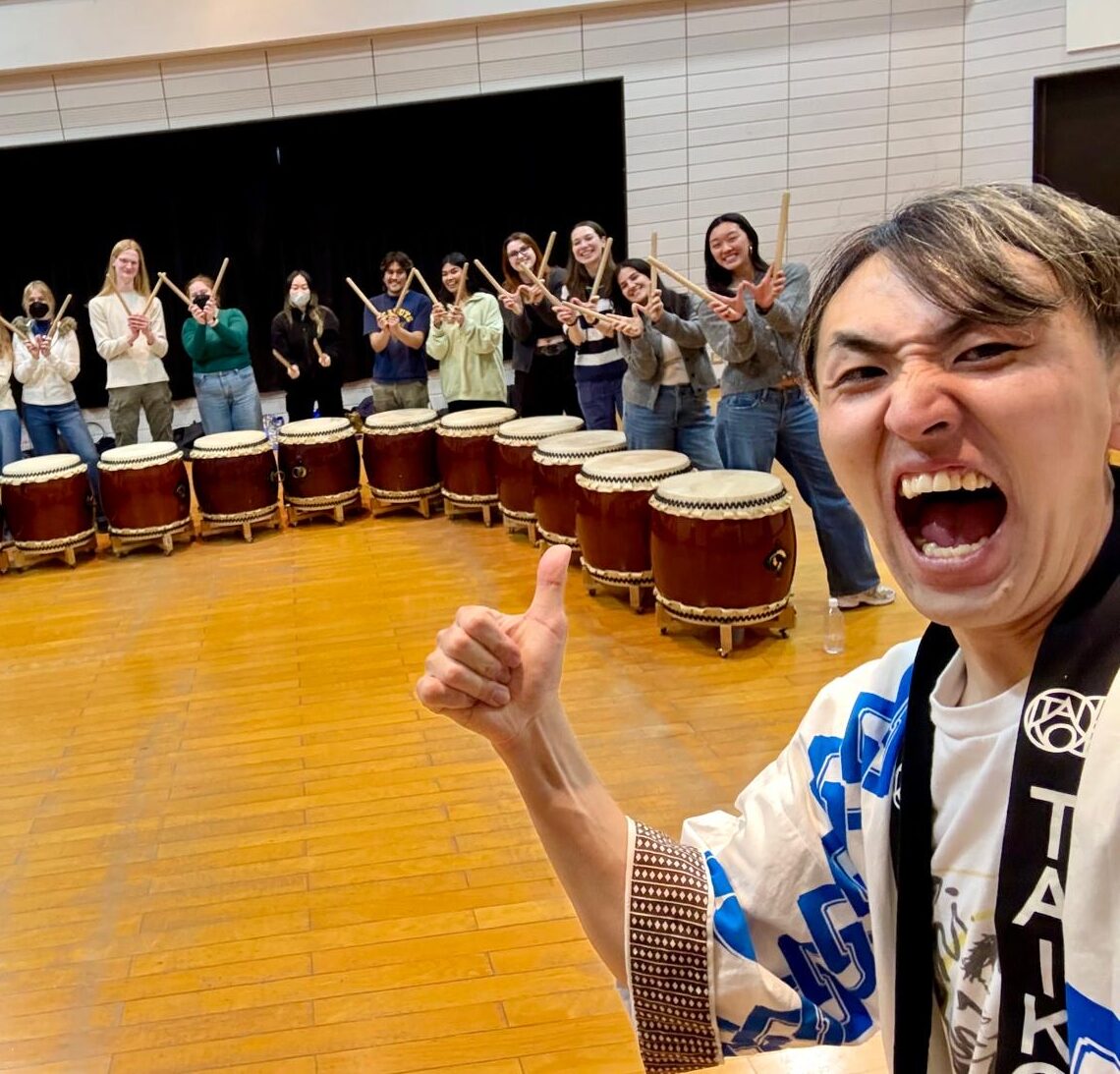 A student makes a "thumbs up" and gestures to a line of classmates behind them. Each classmate stands next to a floor drum and holds up drum sticks to from "W"s.