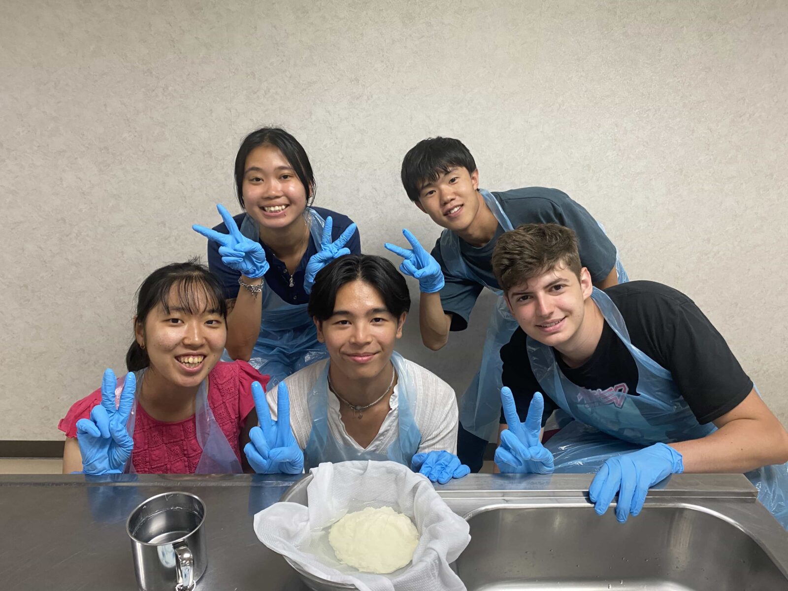 Five students, posing around a bowl that they’re making cheese in, in a large kitchen.