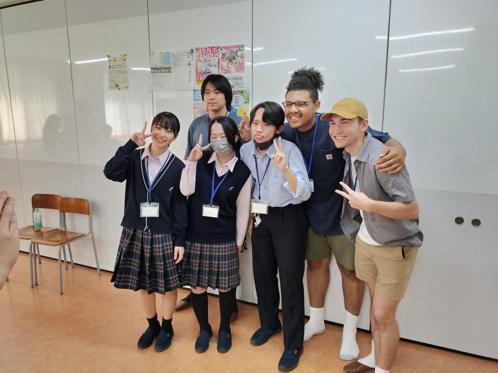 Six students posing as a group, in a classroom, holding up peace signs.