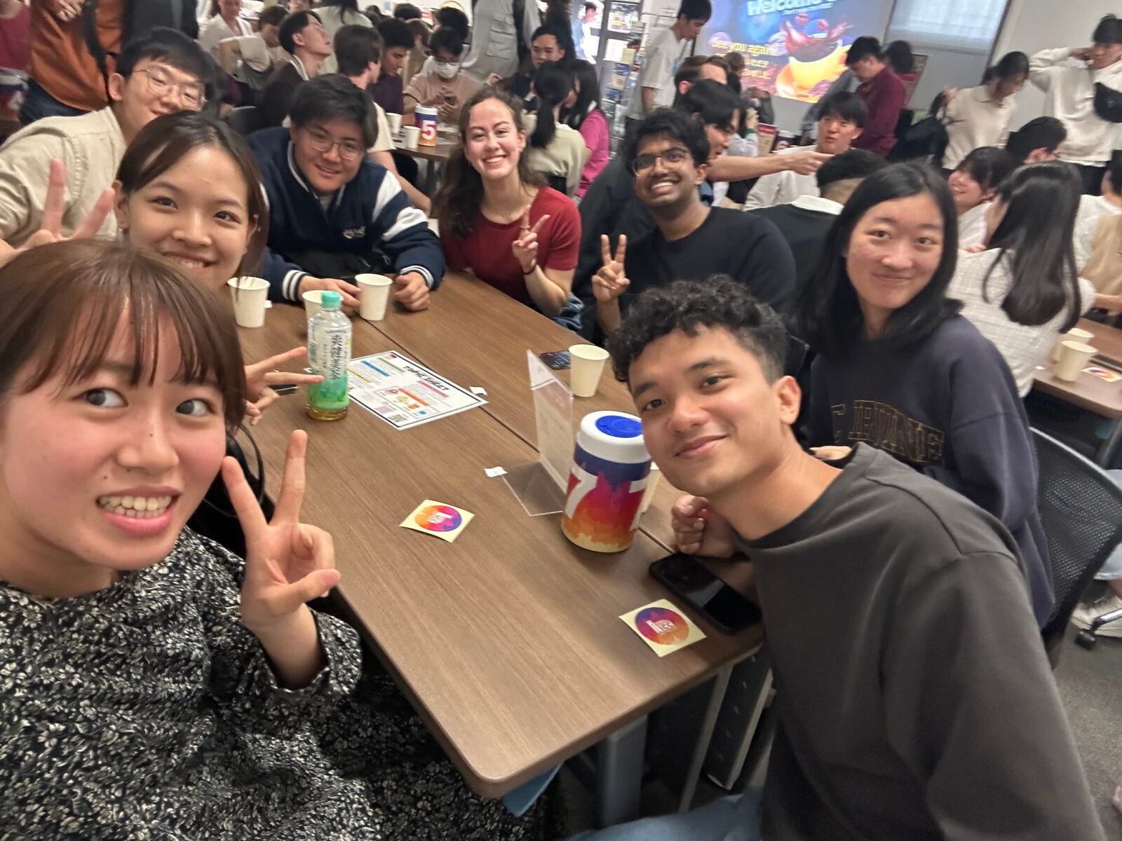 Students sitting around a medium-sized table with drinks, smiling for a selfie.