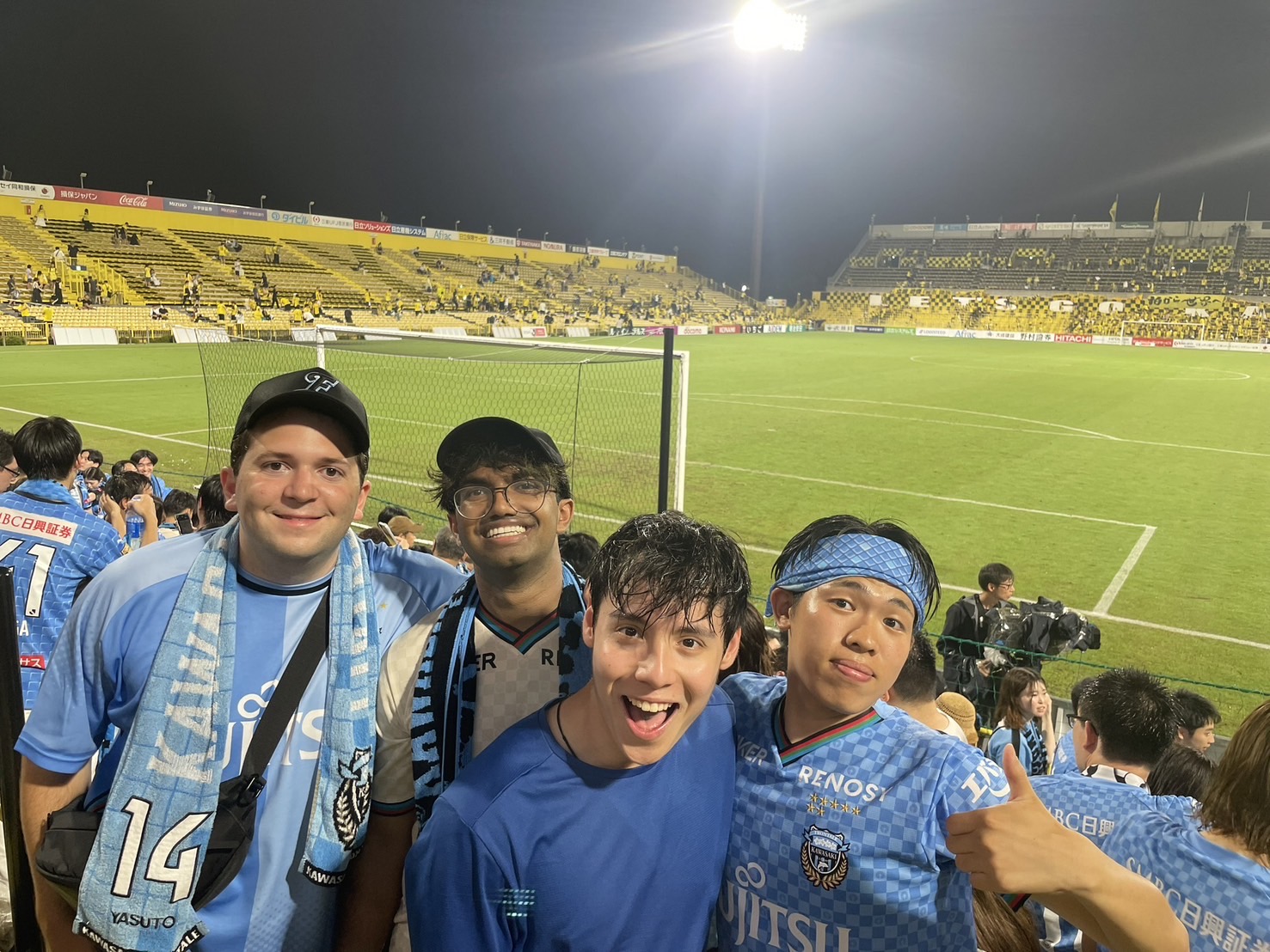 Four students wearing blue soccer jerseys, posing for a picture in a full soccer stadium at night.