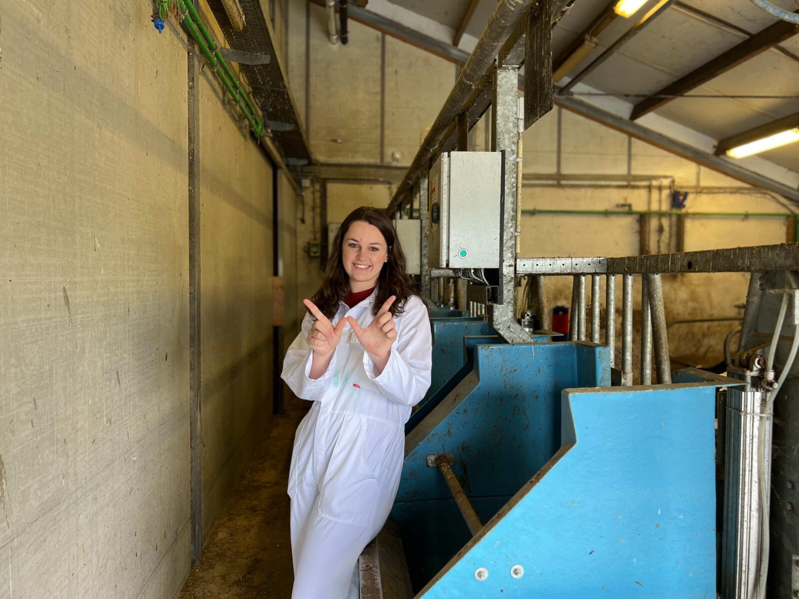 A student holding up a finger W, wearing a white lab coat, standing next to feed bins and other agricultural equipment.