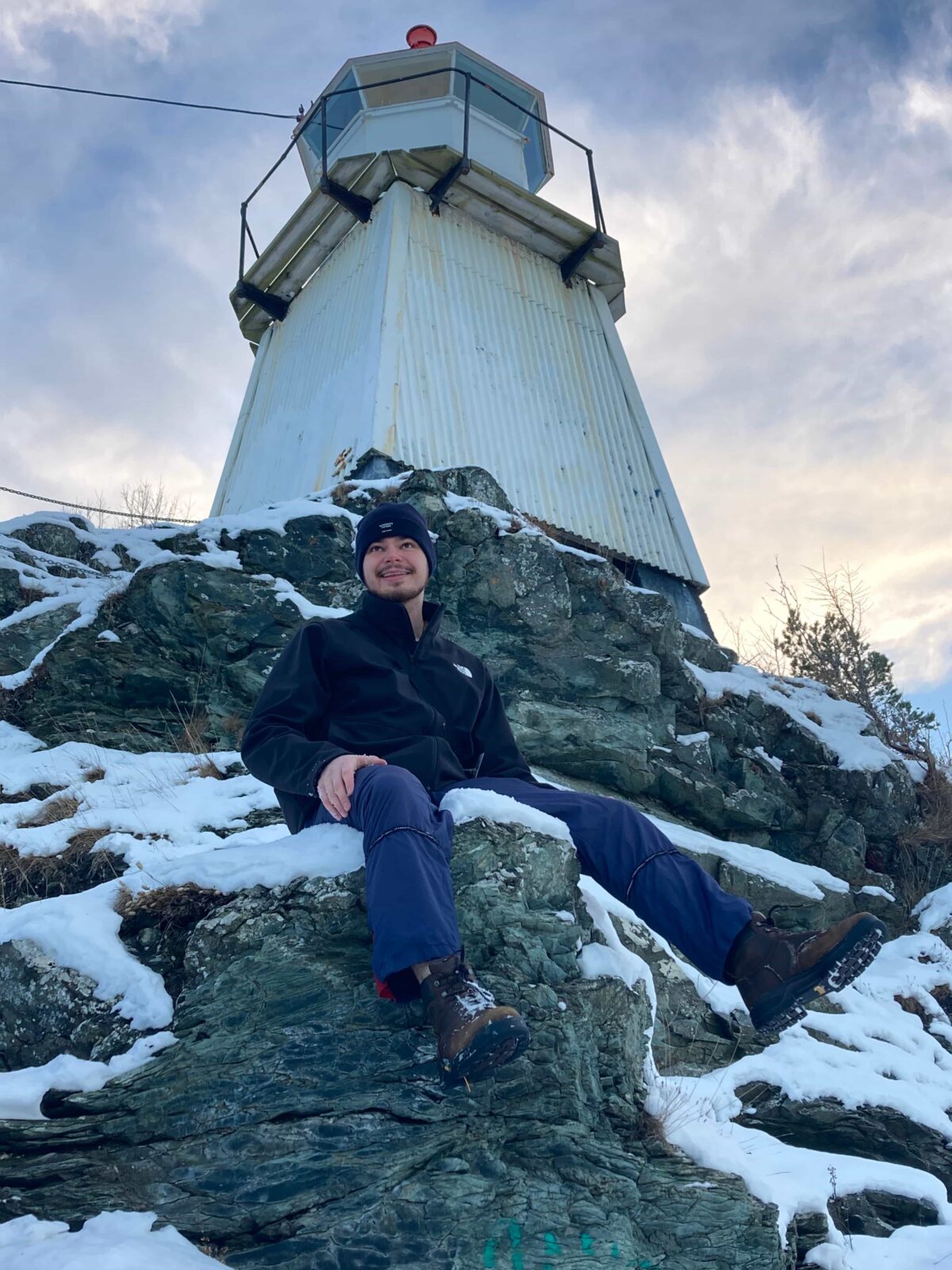 A male student in a blue beanie sits on a snow-covered rock in front of a white, sheet-metal lighthouse.