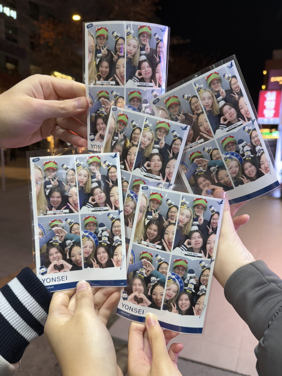 Five hands hold up photobooth photos, each containing four photos of people posing.