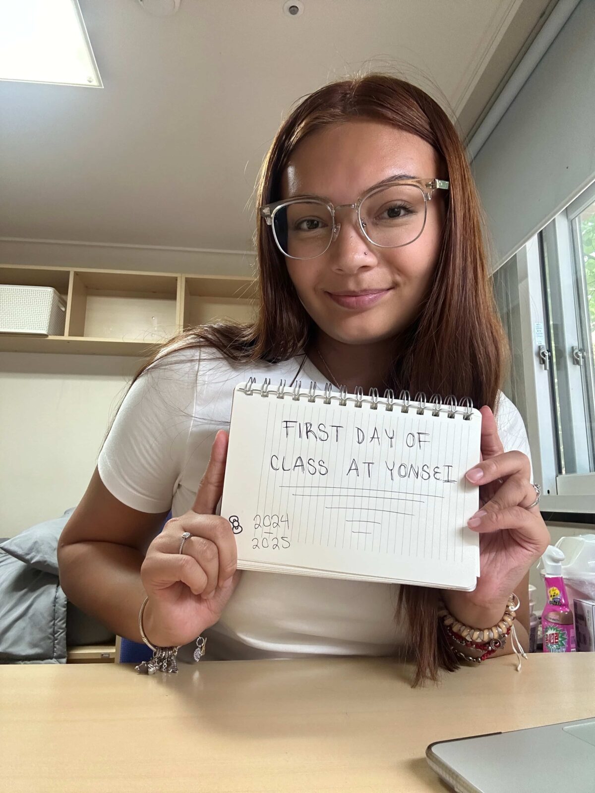A student sitting at a desk, holding up a notebook with the words “First Day of Class at Yonsei”.