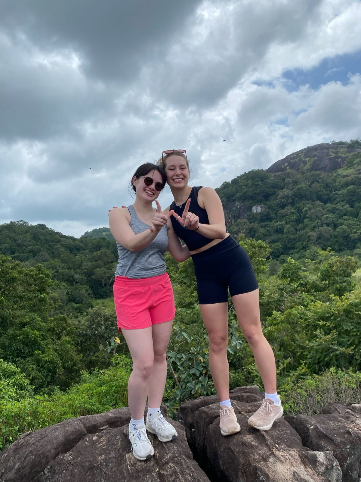 Two students wearing hiking clothes stand on a rock and make a "W" with their fingers. Forested hills and clouds can be seen behind them.
