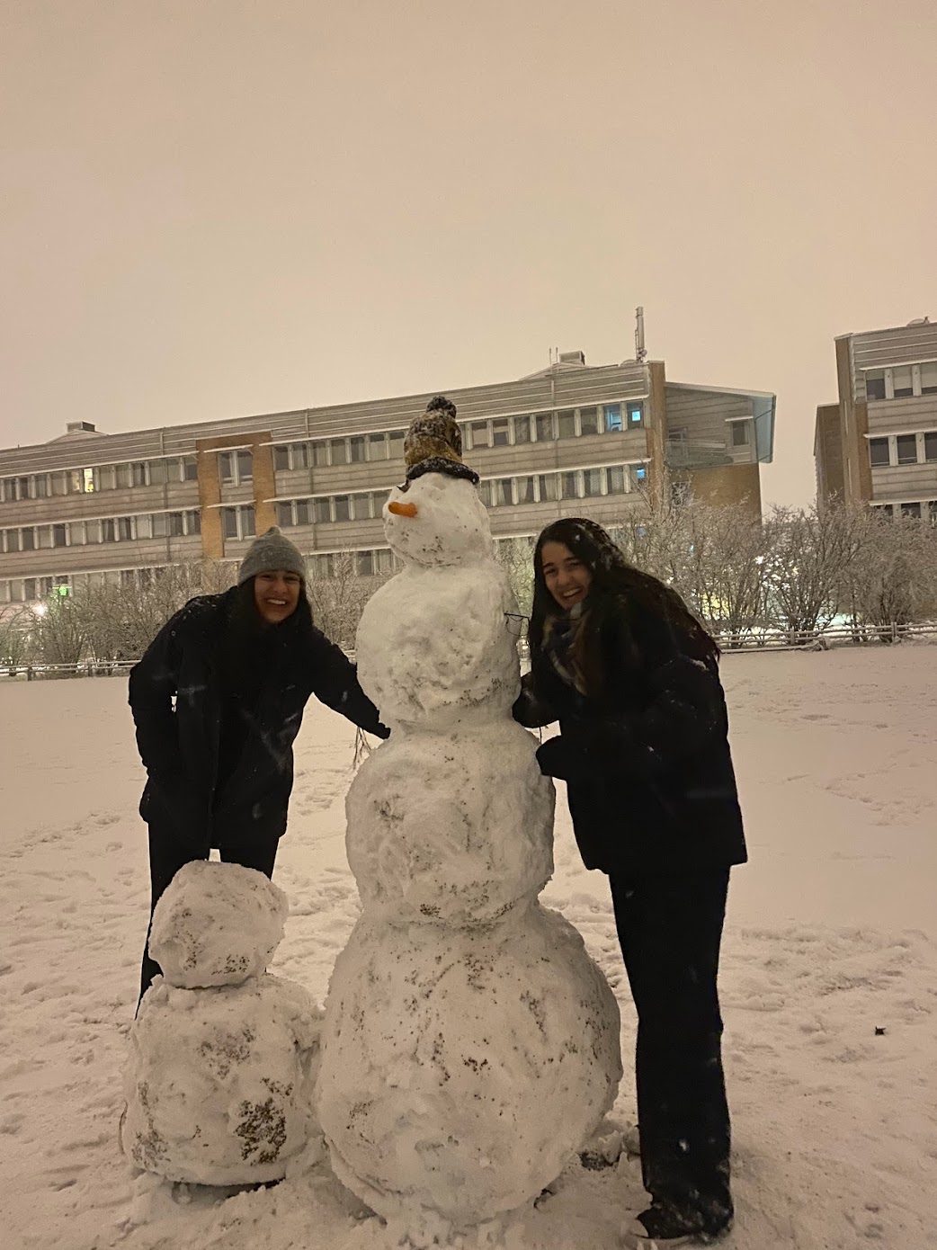 Two students stand on either side of a snowman and smile. The snowman has a carrot nose and is wearing a winter hat. It is taller than the students. There are also two small balls of snow stacked on each other beside it.