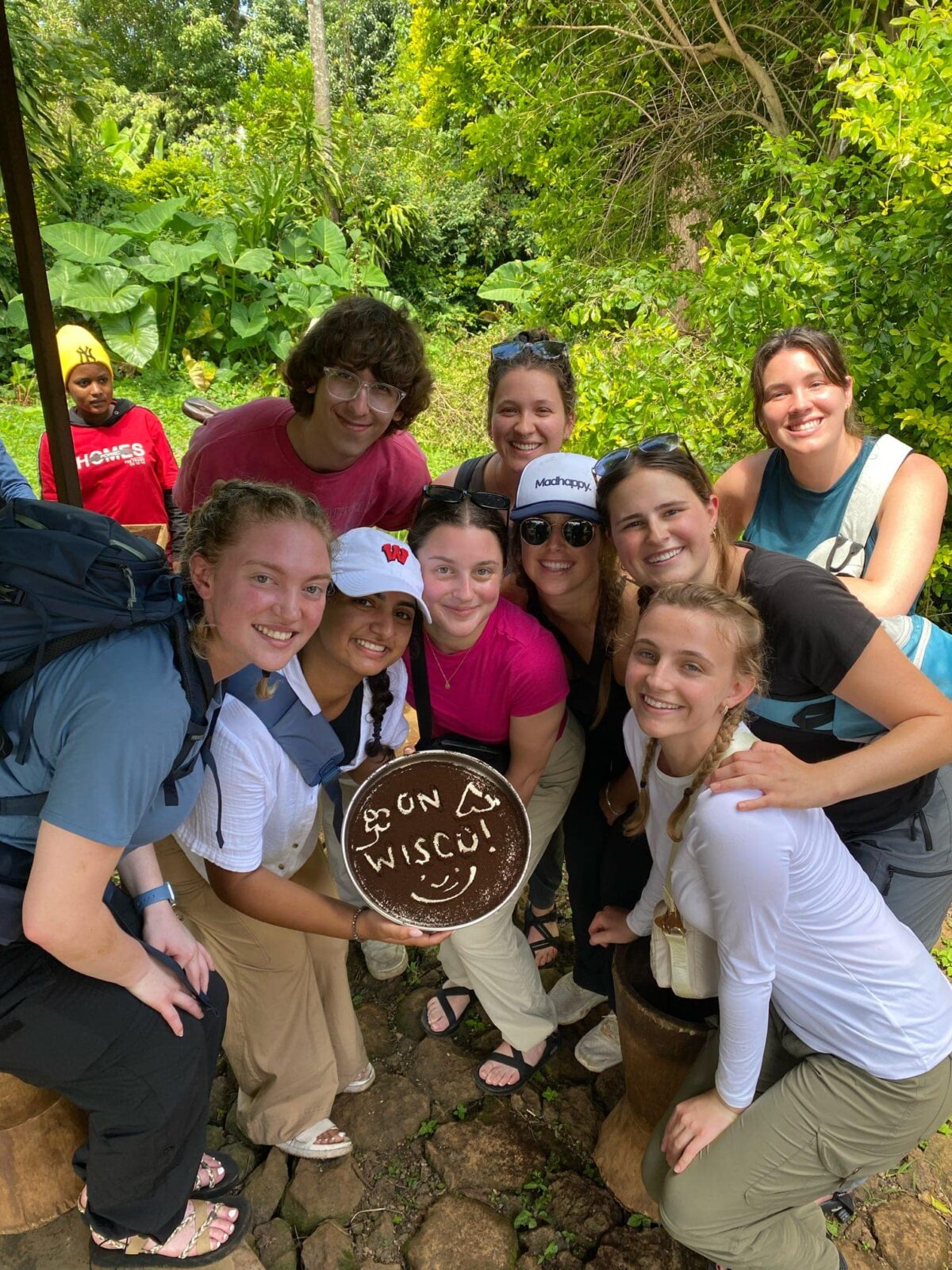 A group of students holding a tray of coffee grounds. The words "On Wisco!" and a smiley face are drawn into the coffee grounds.