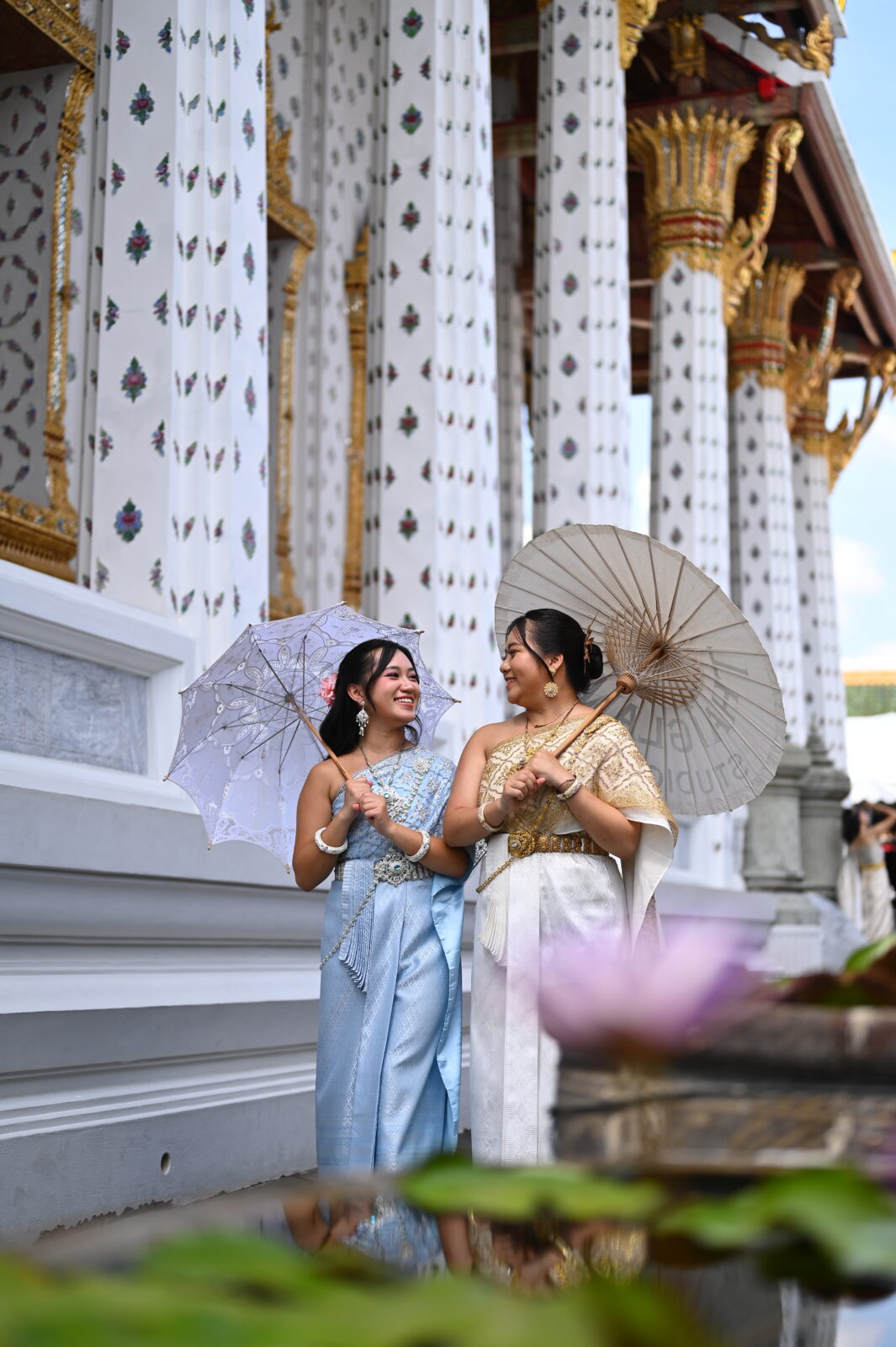 Two women in traditional Thai dresses hold up sun umbrellas in front of a temple with white marble pillars.