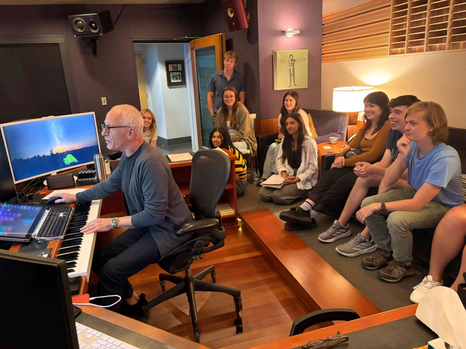 Students sit clustered on a couch watching a man in front of them. The man is siting at a desk with several monitors, a soundboard, and electronic piano. 