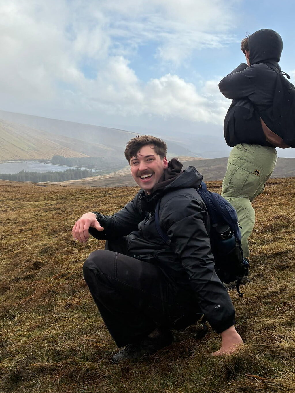 A student wearing a jacket and backpack crouches on a grassy slope while on a hike. Behind him are rolling grassy hills and a lake.