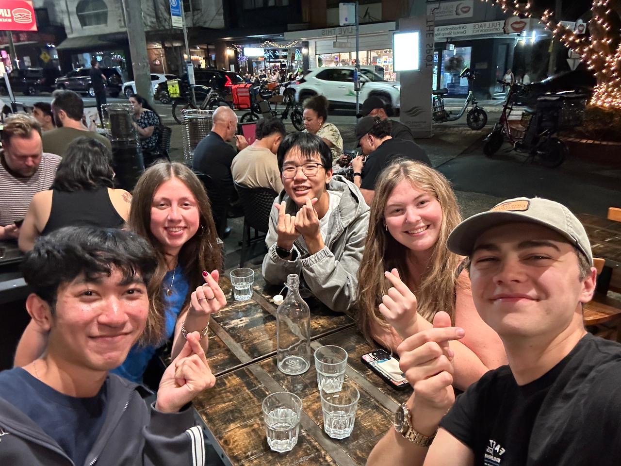 A group of five students, gathered around a restaurant table on a sidewalk, posing for a selfie.