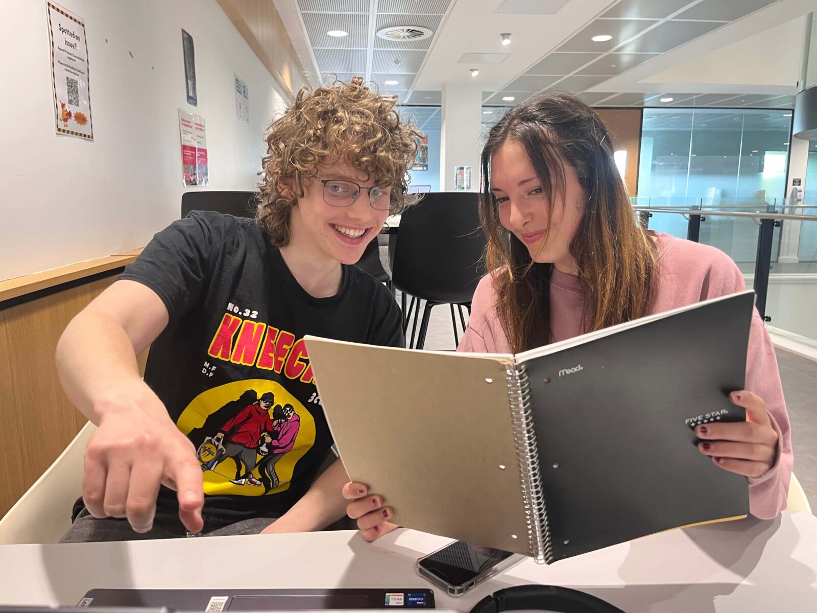 Two students at a table, studying, looking at a computer and notebook.