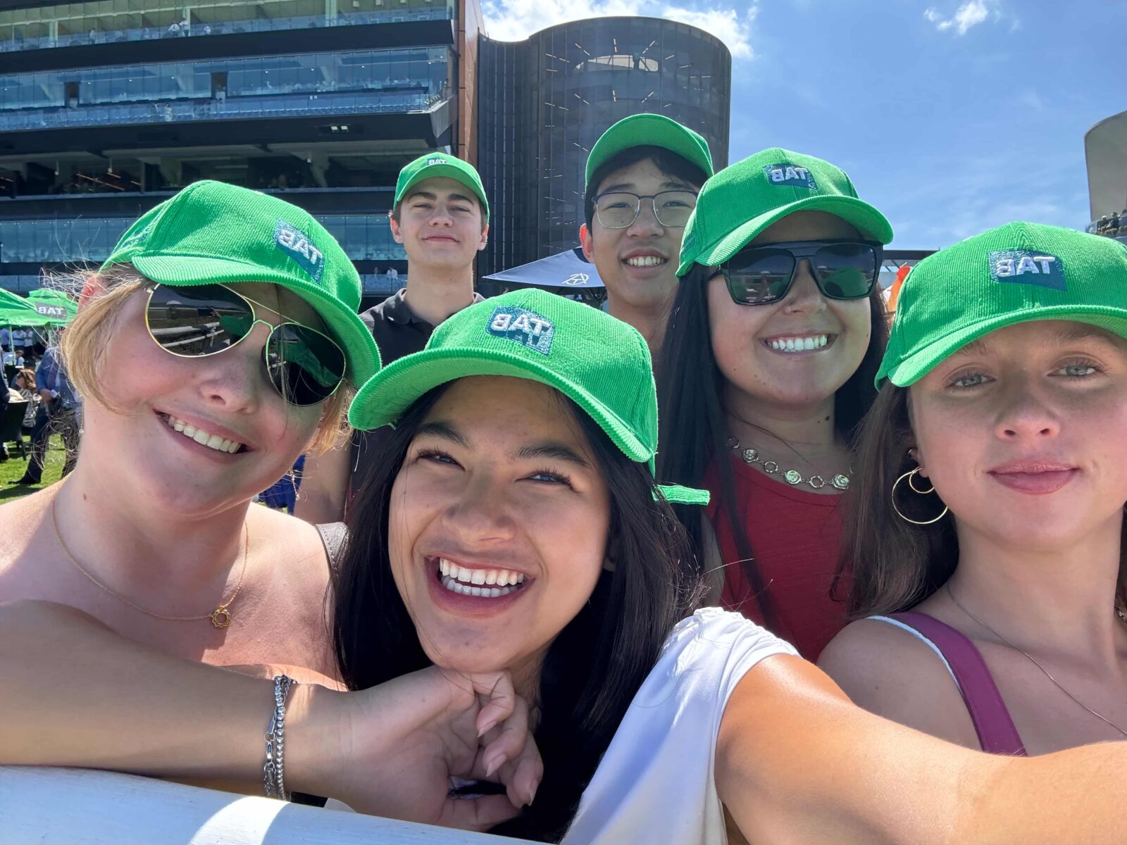 A group of six students wearing bright green baseball hats, standing outside on a sunny day.