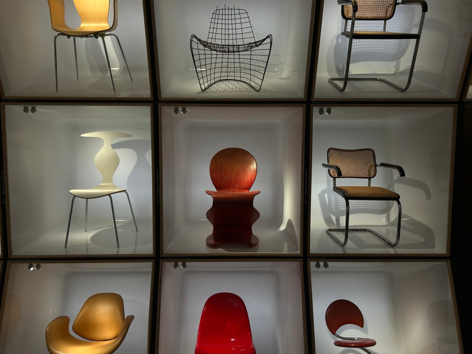 A large glass display case, taking up an entire wall, featuring nine distinct and artistic chairs.