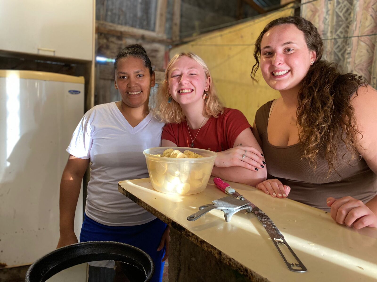 Three individuals posing around a bowl of cut and peeled plantains in a kitchen.