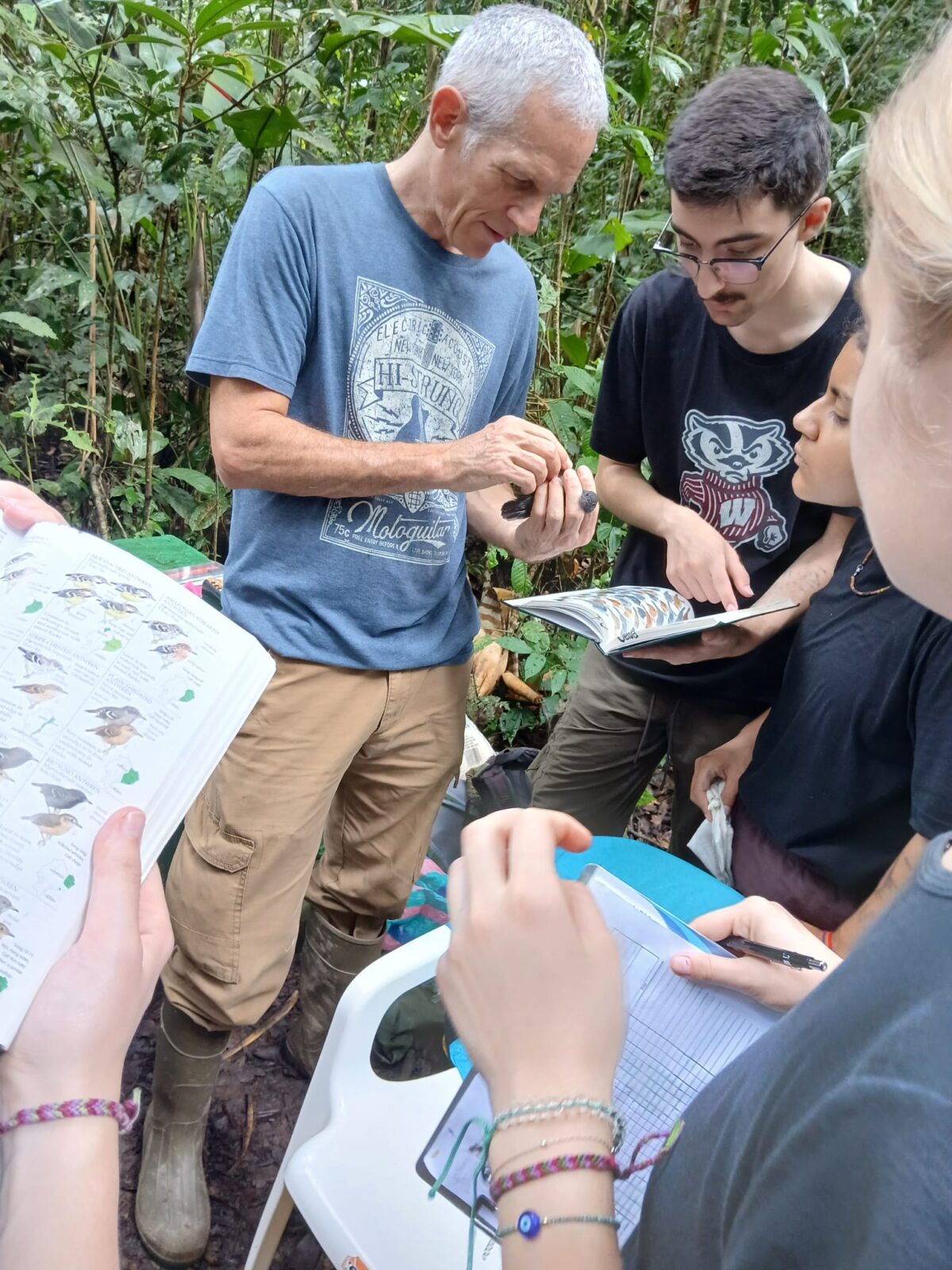 A group of students outdoors, holding notebooks and books, gathered around a professor holding a bird in his hands.