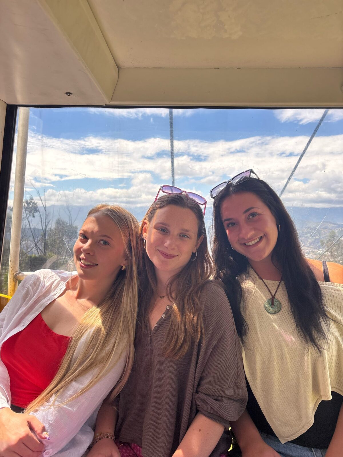 Three students posing in a cable car, with city and mountains visible in the distance behind them.