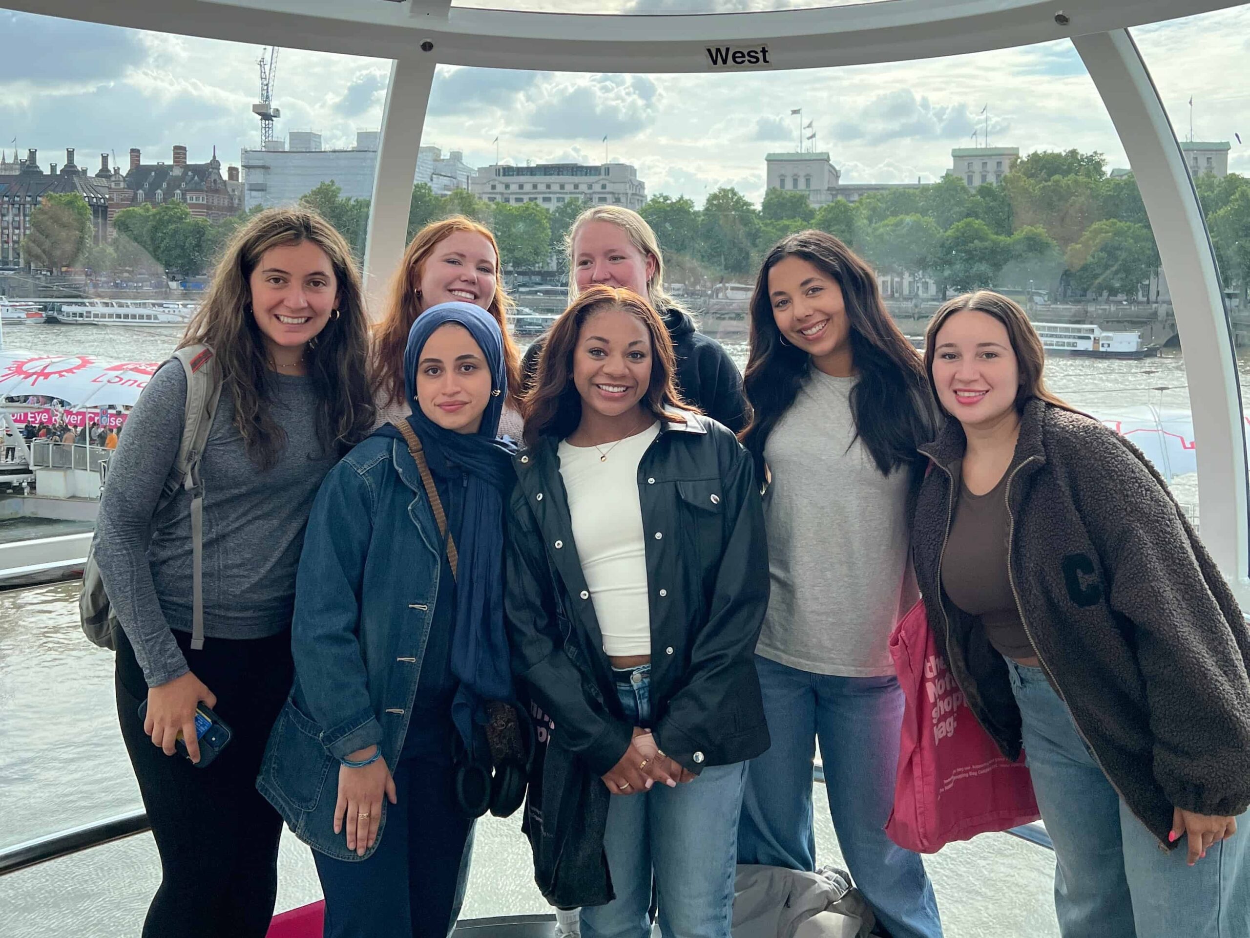Seven students on the London Eye Ferris wheel, in a glass pod, low to the ground. 
