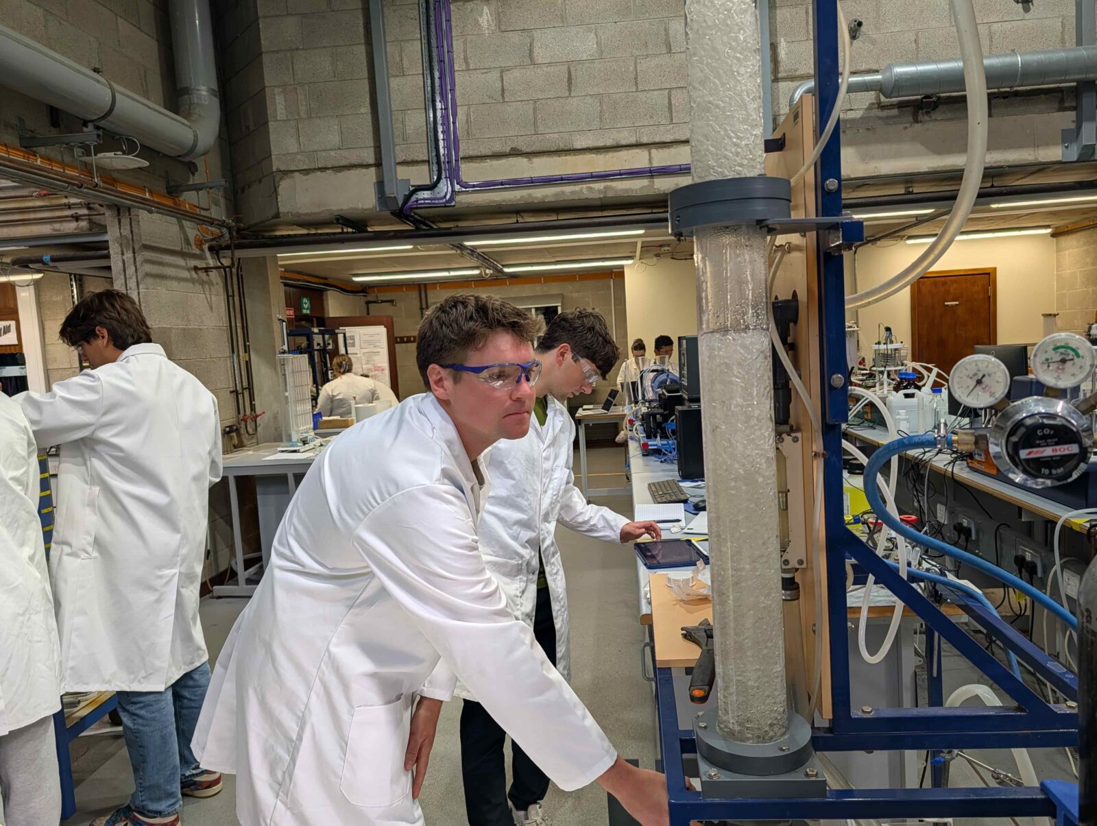 Two students wearing white lab coats look at a large clear vial with what appears to be water with large bubbles in it.