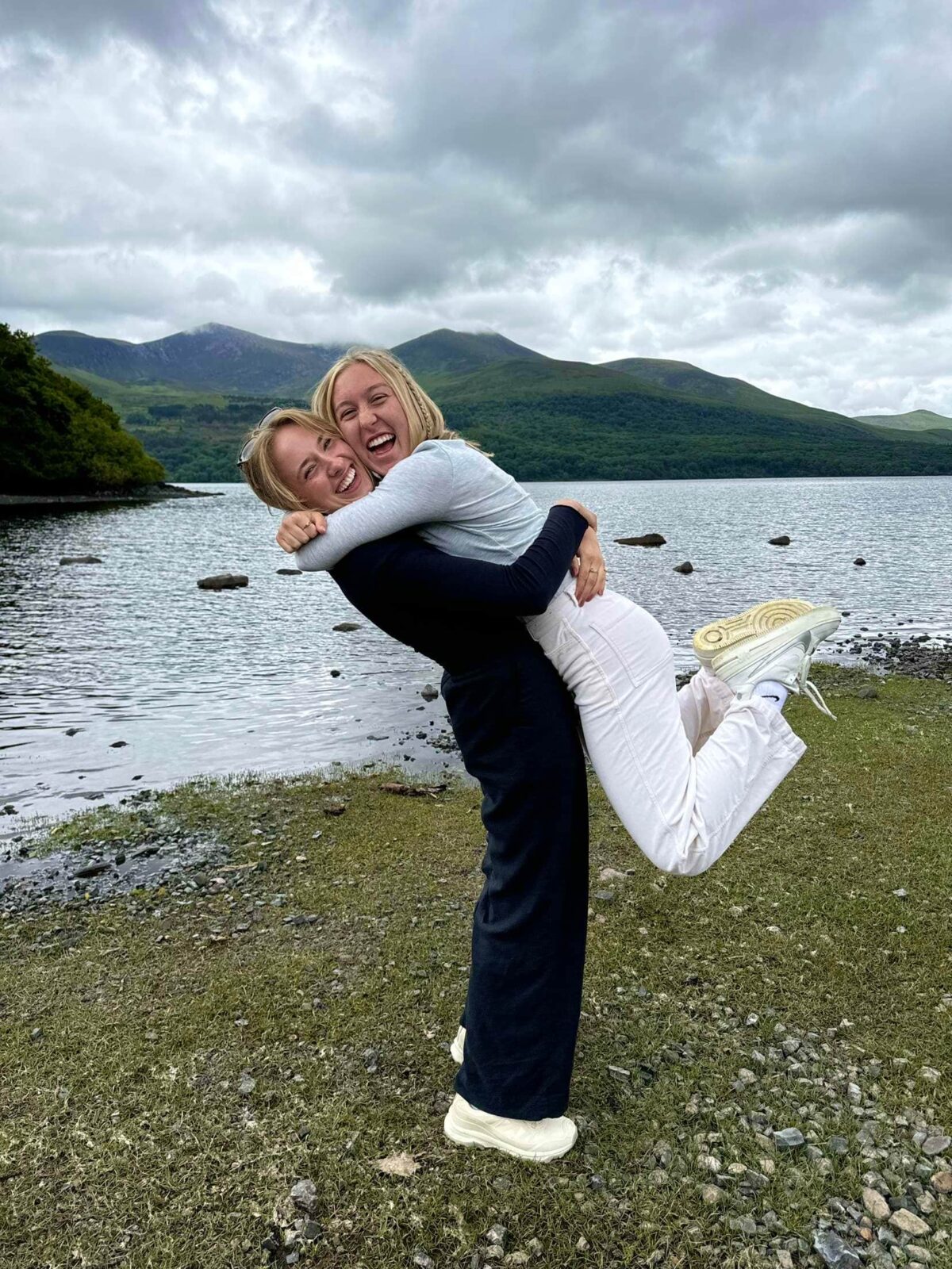 One student lifting up another student while standing on grass sprinkled with rocks in front of a lake surrounded by grassy mountains.