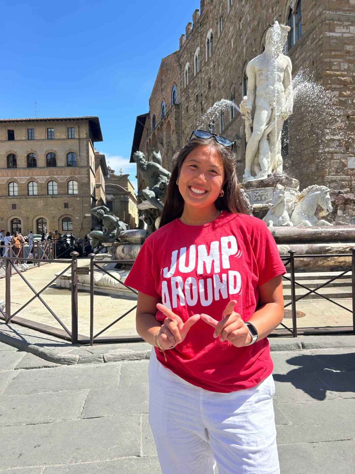 A student wearing a red "Jump Around" shirt holds up her hands in a "W" and stands in front of a statue in a fountain.