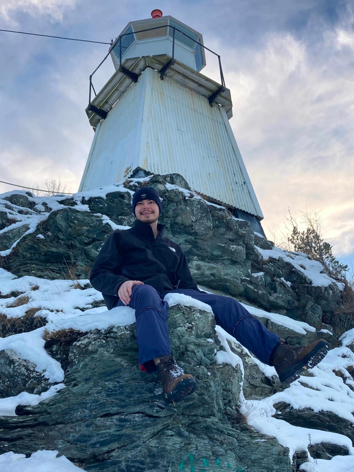 A student in a blue beanie sitting on a snow-covered rock in front of a white, sheet-metal lighthouse.