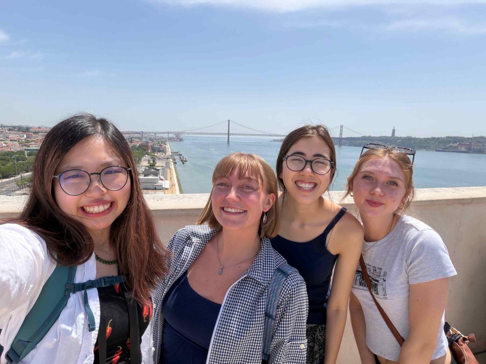 Four students posing for a selfie at a viewpoint over a wide bay, with a large bridge in the background.