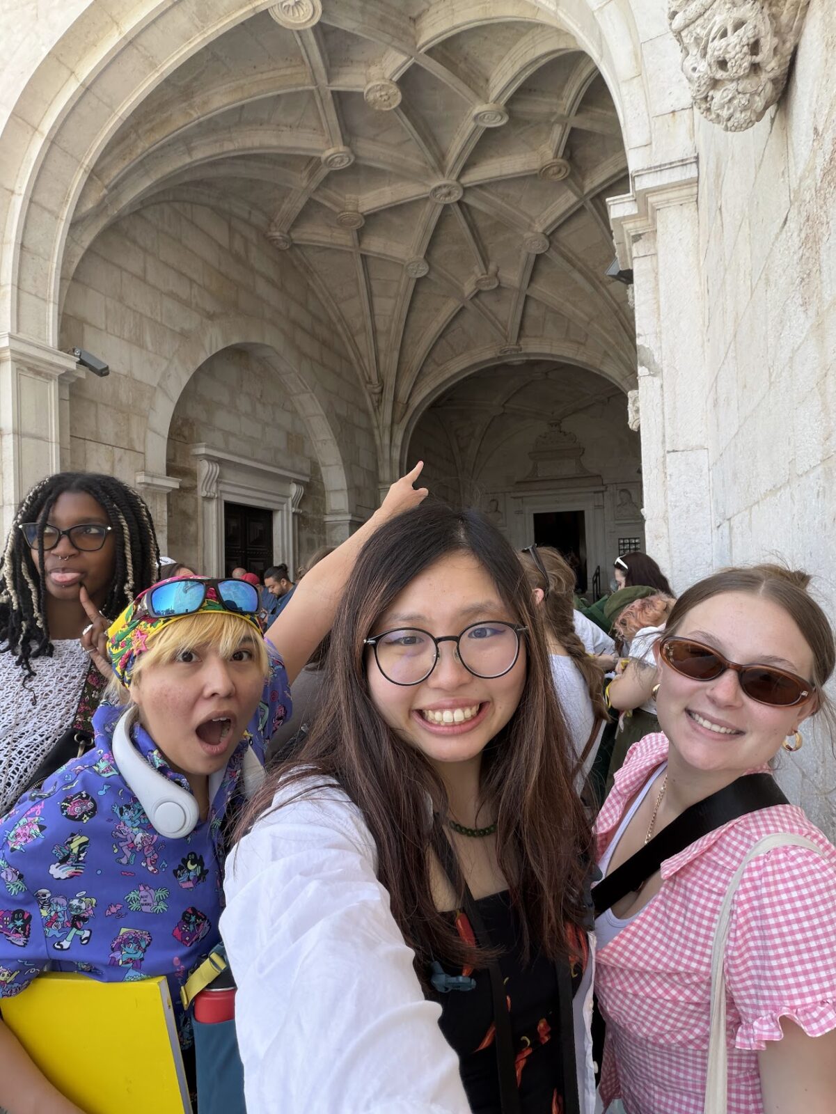 Four students posing for a selfie, smiling and making silly faces underneath an ornate beige semi-circular arched opening.