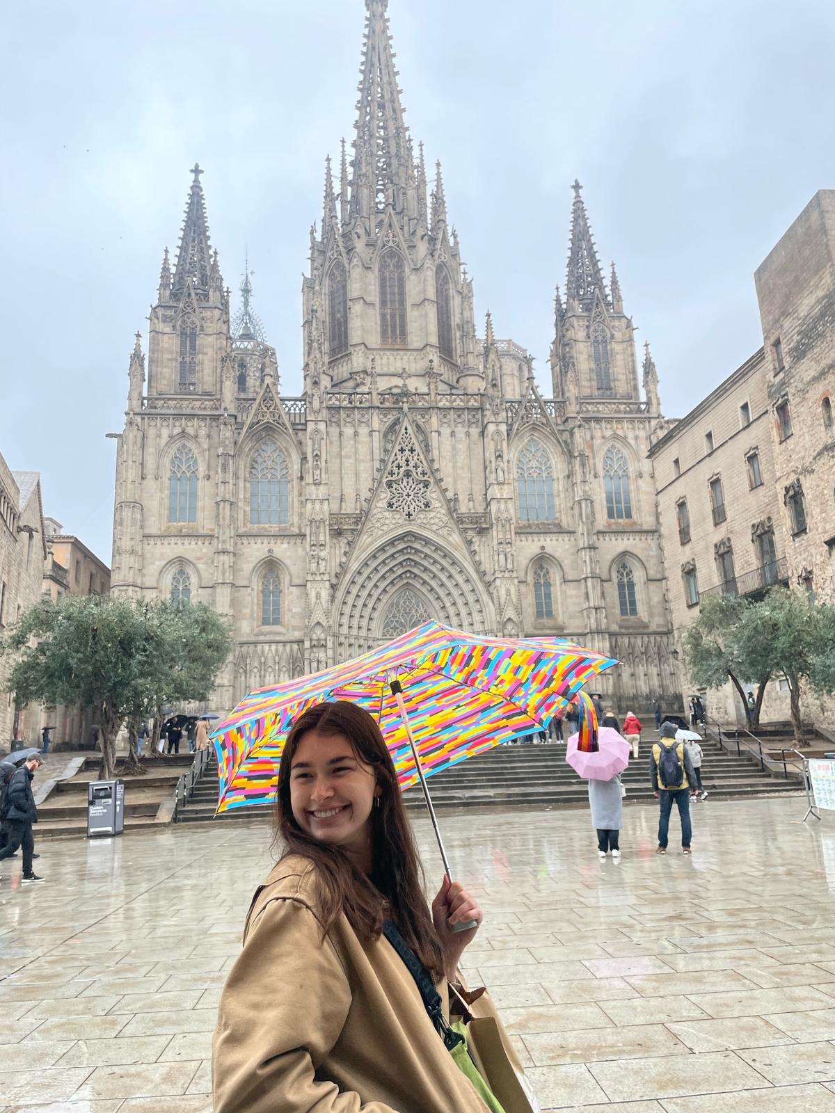 A student holding a multi-color umbrella while smiling in front of the ornate Cathedral of Barcelona.