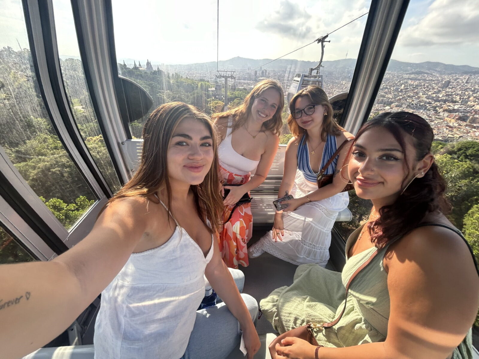 Four students posing for a selfie inside a glass cable car, with buildings and hills visible below and in the distance.