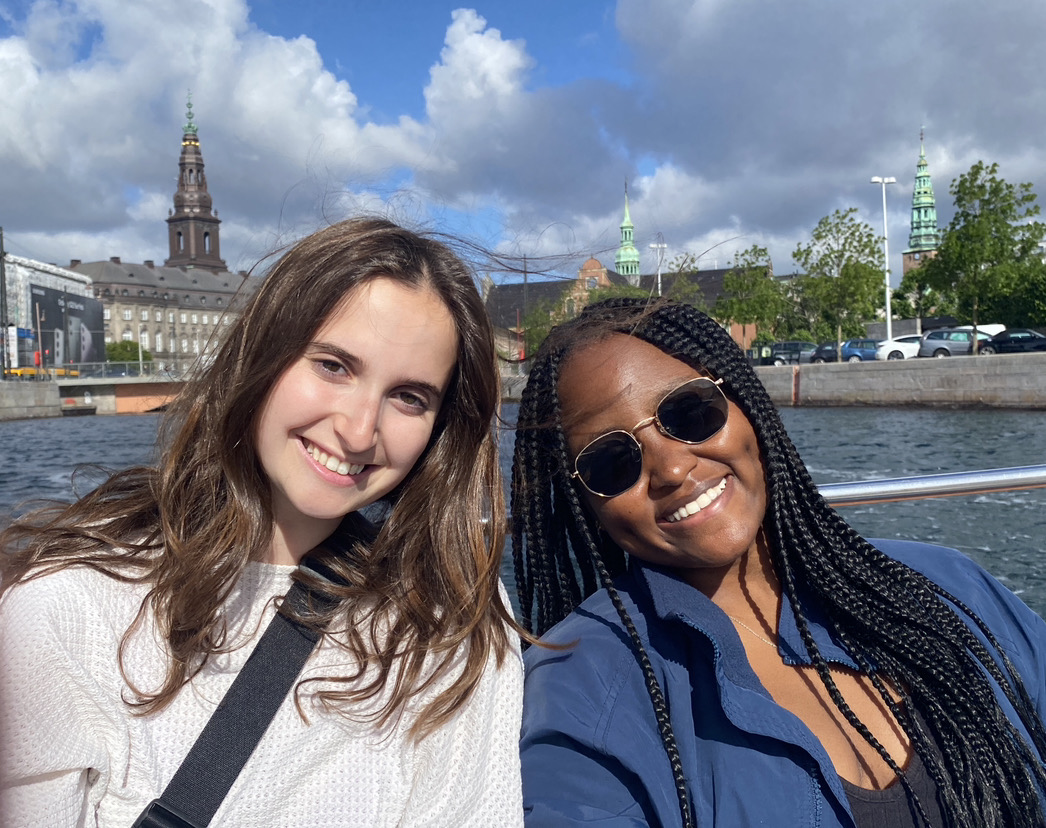 Two students on a boat, smiling, with city buildings and a bridge behind them.