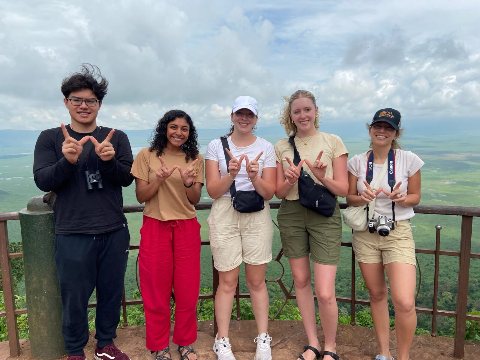 Five students posing at a high overlook, holding up finger Ws, with a wide view of a valley and hills in the distance below them.